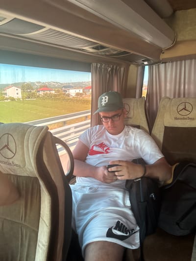 A young man wearing a green baseball cap, glasses, a white t-shirt with a red Nike logo, and white shorts with a black Nike logo sits on a bus. He is looking down at a black mobile phone in his hands. The bus seats have Mercedes-Benz logos. Outside the window, there are green fields, houses, and hills. The scene appears to be in Fushë-Krujë, Albania.