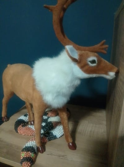 The image displays a close-up, eye-level shot of a stuffed toy reindeer with prominent antlers and a white ruff around its neck. The reindeer is a light brown color, with darker brown accents on its legs and hooves. Its eyes are dark, and its snout is a darker brown. The reindeer is positioned on a wooden surface, leaning its head against a vertical wooden beam. Underneath the reindeer, a beaded snake figurine is coiled. The snake has a colorful pattern of white, black, and orange beads, with green beads for its eyes. The background is a solid, dark blue wall. The lighting appears to be artificial, casting soft shadows. No specific time of day or weather conditions are discernible, nor are any emotions explicitly conveyed by the inanimate objects. There is no visible text in the image. The scene is suggestive of a decorative display, possibly in a home or retail environment, with the toys arranged on a shelf or table.
