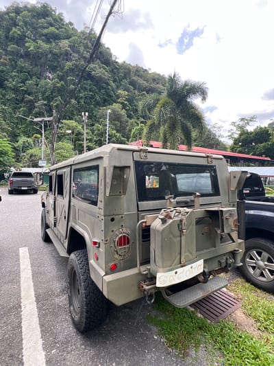 The image shows a military-style vehicle parked on the side of a road. The vehicle is olive green and appears to be a heavy-duty truck or armored personnel carrier. It has large tires, a rugged exterior, and what looks like a spare tire mounted on the back. A metallic step is attached to the side of the vehicle, and a metal grate is visible on the ground next to it. The vehicle is parked on an asphalt road, with a grassy verge to the right.
In the background, a lush, green, densely forested hillside rises steeply. Palm trees are visible among the trees. The sky is overcast, suggesting cloudy weather. Electrical wires and poles are strung along the side of the road, indicating a populated area. Another vehicle, a dark-colored pickup truck, is parked behind the military vehicle, and a glimpse of another vehicle is visible through the windows of the military vehicle.
The scene appears to be in a rural or semi-rural setting, possibly a tropical or subtropical region given the vegetation. The lighting suggests it could be daytime, though the overcast sky creates a diffused light. There is no clear indication of any activity taking place, as the vehicles are stationary.
There is some visible text on a white plate on the rear of the military vehicle, but it is not clearly legible. The overall impression is one of a sturdy, utilitarian vehicle parked in a natural, verdant environment.