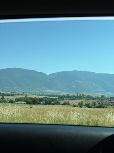 This image captures a scenic view from a window, looking out onto a rural landscape. The foreground shows the dark interior of a vehicle, framing the outdoor scene. Beyond the window, a field of tall, dry grass stretches towards a middle ground dotted with trees and some scattered buildings. In the distance, a range of rolling, tree-covered mountains forms the backdrop under a clear blue sky. The overall impression is one of a tranquil, sunlit day in a village setting, likely in the countryside surrounding Orizari, Bulgaria.