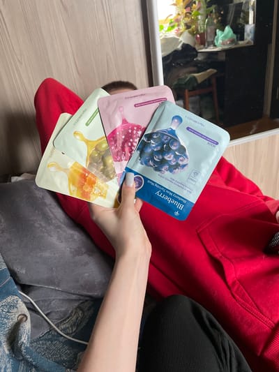 A woman holds four facial mask packets. The top mask is blue with an image of blueberries, labeled "Blueberry Blueberry Mois Turizing Mask". Underneath is a pink packet with a pomegranate design, described as "POMEGRANATE MOISTURIZING MASK". Two light green packets, each with depictions of olives and honey, are located below. All packets are labeled "30ge1.00fl.OZ.U.S". The woman wears a red hoodie and black top. The background includes a mirror reflection and part of a gray garment with blue details. The location is not Ulukulevo, Russia.