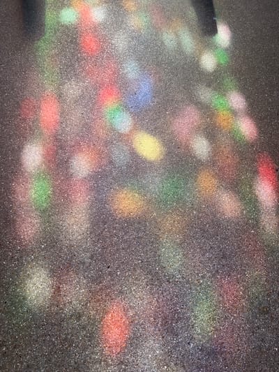 The image shows a section of asphalt pavement on which colorful light patterns are cast. These patterns appear as blurred, elongated shapes of various hues, including red, pink, green, yellow, and blue. The asphalt itself is a dark gray material speckled with lighter aggregate. The light suggests a source such as stained glass or a prism, creating a dappled effect on the ground. The scene is devoid of people or specific objects beyond the pavement and the light. There is no discernible context that would place this image in the city of Rambouillet, France.