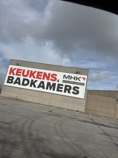 The image displays the exterior of a commercial building and an adjacent asphalt parking lot in Roeselare, Belgium. The building features a light-grey concrete panel facade. A large white sign, approximately two-thirds the height of the visible building section, is affixed to the upper facade. The sign displays text in red and black capital letters: "KEUKENS" in red, followed by "&" and "MHK" in black. Below this, "BADKAMERS" is prominently displayed in large black letters. To the right, a smaller text block reads "KITCHEN EXPERT" in black. Two dark-colored light fixtures are mounted on the facade directly above the sign.
The foreground consists of a dark asphalt parking lot with multiple faint white parking space lines. Numerous dark, curved tire tracks are visible on the asphalt surface. The sky above is largely covered by grey, overcast clouds, with some lighter blue sky visible and faint white contrails present. A dark, blurred object, consistent with a vehicle's interior frame, partially obscures the top edge of the image. No individuals or other vehicles are present in the scene.