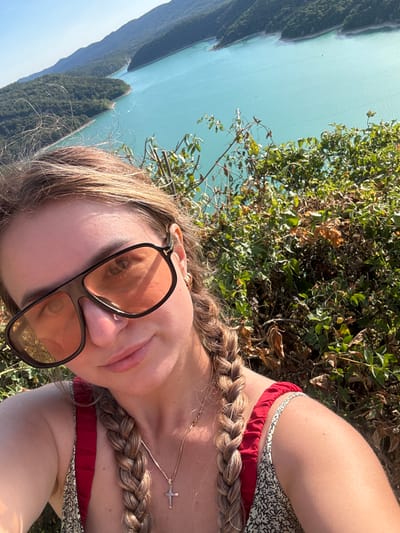 A young woman with blonde hair styled in two braids is taking a selfie outdoors, with a vibrant turquoise lake and tree-covered mountains in the background. She is wearing large, dark-rimmed sunglasses with orange lenses, a red and white patterned top with red ruffled straps, and a gold necklace with a cross pendant. The location appears to be the Lac de Vouglans, near the town of Moirans-en-Montagne, France, a popular tourist destination known for its scenic beauty and recreational activities. The sun is shining brightly, casting shadows and highlighting the lush green foliage around her.