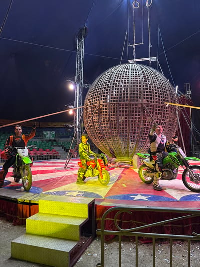 The image shows a "Globe of Death" circus act taking place in Tejalpa, Mexico. Three motorcyclists are riding inside a large, spherical steel cage. The cage is suspended by cables from above. The platform surrounding the globe is painted with an American flag design. A short set of yellow-painted stairs leads up to the platform. The backdrop is a dark blue tent. Spectators are visible in the background. One of the motorcyclists is standing and waving.