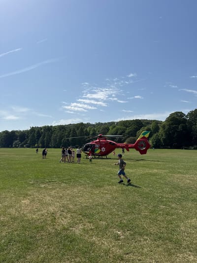 A red air ambulance helicopter is on a grassy field next to a line of trees under a blue sky with some clouds. Several people are standing near the helicopter. One individual is closer, in a running stance. The grass appears to be a playing field. The lettering on the helicopter reads "AIR AMBULANCE". The helicopter registration is visible as G-TVLY. Considering the location, the helicopter likely belongs to the Thames Valley Air Ambulance. Given this context, the location of the scene is likely High Wycombe, United Kingdom.