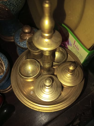 A vintage brass betel nut set, likely from Southeast Asia, features a central pedestal with a knob-like finial. Around its base are several small, round containers, some with lids, arranged on a circular tray with raised edges. The set appears to be tarnished, and a green box with an "Iris" label sits next to it. Nearby are small, decorated porcelain or ceramic containers. The overall scene suggests an antique or curio display.