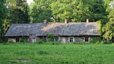 The image shows a long, low-lying stone and brick building with a corrugated metal roof in a rural setting. The building appears to be an old, possibly abandoned structure. It is surrounded by lush green grass and vegetation, with a dense forest of trees in the background. Several windows are visible on the facade of the building, some with glass and others appearing broken or boarded up. Chimneys protrude from the roof. Vines are growing on the sides of the building, indicating a lack of recent maintenance. The overall impression is one of disrepair and a natural, overgrown environment. The location is identified as Daudas, Latvia.