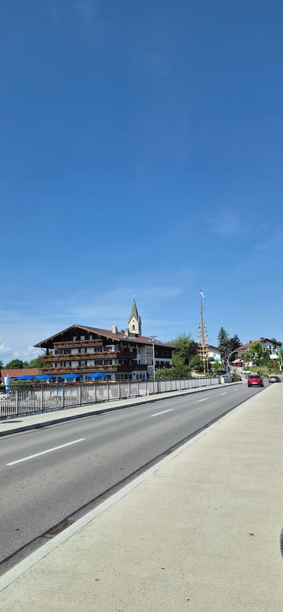 The image captures a scene in Seeon-Seebruck, Germany, dominated by a clear blue sky. In the mid-ground, a large, traditional Bavarian-style building with multiple balconies adorned with flowers is visible. Blue umbrellas are set up in front of the building, suggesting an outdoor seating area. To the right of this building, a tall pole with a flag and decorative elements stands prominently. Further in the background, a church steeple rises above the other structures, and several other buildings and trees are present. A street with a white dividing line runs across the foreground, with a light-colored sidewalk to its right. Two cars are on the road, one red and one dark-colored, moving away from the viewer. A white metal fence lines the area between the building and the road. The overall environment appears to be a sunny day in a town or village setting, with elements of architecture, nature, and transportation.