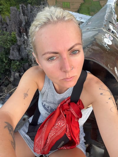 A woman with blonde hair and blue eyes takes a selfie outdoors. She is wearing a grey tank top and grey shorts, with a red bandana tied around her waist. She has several tattoos on her arms and a nose ring. Behind her, there are jagged grey rock formations and lush green vegetation. To her right, a weathered, metallic structure with stickers is visible, suggesting she is near a monument or a piece of machinery. The background shows a landscape of green fields, indicating a rural or semi-rural setting. The scene appears to be located in the vicinity of the Pha Ngum viewpoint in Ban Phon Sai, Laos, known for its dramatic limestone karsts and expansive vistas.