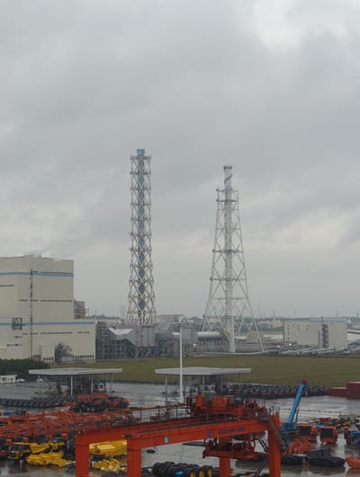 This image depicts an industrial complex in Tōkai, Japan, under an overcast sky. The scene is dominated by two large, lattice-style towers, likely smokestacks or cooling towers, that rise prominently against the cloudy atmosphere. In the foreground, a large orange gantry crane is visible, along with numerous construction vehicles such as excavators and other heavy machinery parked in rows. The ground appears wet, suggesting recent rain.

The industrial setting is further emphasized by various buildings, pipes, and power lines. The presence of the construction equipment suggests ongoing work or a storage area for machinery. The overall mood is somber due to the cloudy weather. No people are clearly discernible in the image, and no text is legible. The time of day appears to be daytime, given the ambient light.