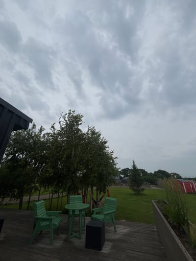 The image depicts an outdoor seating area with a wooden deck and three green chairs and a green table. Behind the seating area are several tall trees, and beyond that, a grassy field with scattered buildings and a red structure. The sky is overcast with grey clouds. The scene is located in Tavrichanka, Russia. There are no people visible in the image. The overall atmosphere is somber due to the cloudy sky.
