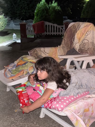 In Enevo, Bulgaria, a young girl with dark, wavy hair lies on a white plastic lounge chair outdoors at night. She is covered with blankets, one pink with white polka dots and another light blue with a star pattern. She is holding a red bag of chips, labeled in Cyrillic script, with her left hand. The background shows a white fence, green bushes, and a large water bottle. The scene appears to be a backyard or patio area.