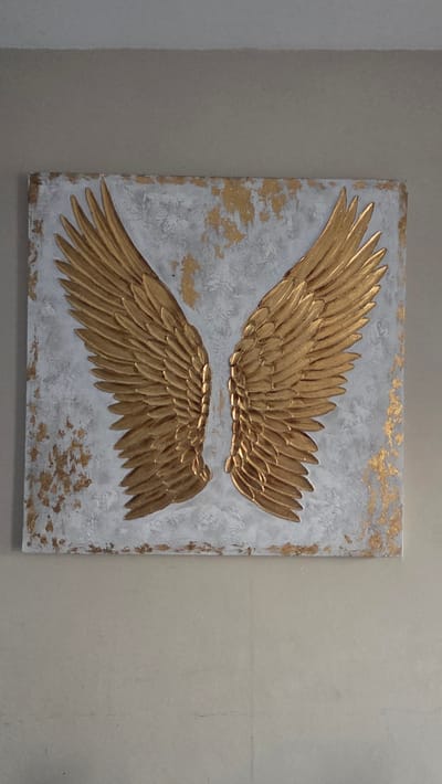 The image shows a square painting of angel wings. The wings are gold and appear to be textured, with individual feathers clearly defined. They are mounted on a white background with flecks of gold leaf scattered across it, creating a distressed or abstract effect. The painting is hung on a plain, neutral-colored wall. The lighting is soft, casting subtle shadows that highlight the relief of the wings. The context of the city of Macé, France is not discernible from the visual information in the image.