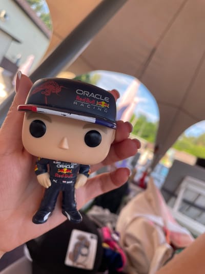 A close-up shot shows a hand holding a Funko Pop toy of a Formula 1 driver. The toy is wearing a dark blue racing suit with red and yellow accents, featuring the "Oracle" and "Red Bull Racing" logos, as well as "Max" written on the leg. The driver's helmet is black with red and white trim and also displays "Oracle Red Bull Racing" and a bull graphic. The toy is held at an angle, obscuring the face. The background is blurred, suggesting an outdoor setting with a pale canopy overhead and hints of greenery and sunlight. The focus is on the toy and the hand holding it. The lighting suggests it is daytime, possibly sunny. The context provided indicates the location is Zazymia, Ukraine, though this is not visually discernible from the image itself.
