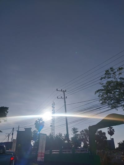 The media file captures an outdoor scene in Macege, Indonesia, likely during the daytime. The image is dominated by a vast, clear blue sky with a bright sun creating a strong lens flare. Overhead, a complex network of electrical wires crisscrosses the frame, supported by utility poles. Lush green foliage from trees is visible in the upper left and right portions of the image, suggesting a somewhat tropical environment. In the background, silhouetted against the bright sky, are buildings and a palm tree, indicating an urban or semi-urban setting. A tall communication tower can be seen in the middle ground. Some text is visible on signs in the background, including "S2 050" and what appears to be Indonesian script on other signs. The strong sunlight and clear sky suggest fair weather. The composition, with the wires and sun creating a diagonal line, gives a sense of depth and perspective.