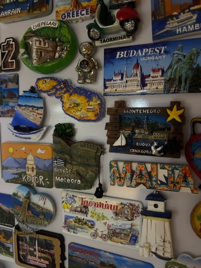 A tight, eye-level photograph captures a densely arranged collection of approximately twenty-two distinct tourist souvenir magnets affixed to a smooth, glossy white vertical surface, characteristic of a refrigerator door. The surface displays minor reflections from an unseen light source.
The magnets vary in shape, size, and three-dimensionality, depicting international landmarks, cityscapes, and country outlines. Prominently visible magnets include: a round, green relief magnet labeled "CHEPELARE" showing a traditional building; a silver teddy bear magnet featuring a red heart stating "I LOVE VIENNA"; a rectangular magnet illustrating the Hungarian Parliament Building and a statue, labeled "BUDAPEST HUNGARY"; a map-shaped magnet of "MALTA"; a rustic-framed magnet for "MONTENEGRO" displaying a coastal town scene with a small white sailboat; a relief magnet for "Greece Meteora" showing clifftop monasteries and the Greek flag; a rectangular magnet for "KOTOR" depicting a bay and a church spire; colorful patterned letter magnets spelling "MALTA"; and a white lighthouse-shaped magnet. Other visible magnets feature locations such as "BAHRAIN," "GREECE," "TAORMINA" (twice, one with a scooter, one a panoramic card), "HAMBURG" (twice), "SKOPJE," "MACEDONIA" (with "Ohrid Lake"), and "RAPPEN."
No individuals are present, and no actions or interactions are depicted beyond the static display. The immediate environment is limited to the white surface holding the magnets. The image itself provides no visual information to confirm its specific location in the city of Mosomishte, Bulgaria, although one magnet is identified as from Chepelare, Bulgaria.