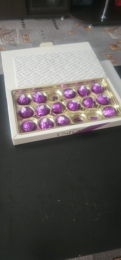The image shows an open box of chocolates. The chocolates are individually wrapped in purple foil and are arranged in a plastic tray within a white cardboard box. The lid of the box is slightly ajar and displays a pattern of repeating text and symbols. The box itself has the brand name "ESFERO" printed on the front edge. The chocolates appear to be spherical. The box is placed on a dark, textured surface that has some scuff marks and light-colored speckles. The background shows a patterned carpet and furniture. The image does not contain information about the city of Vesele, Ukraine.