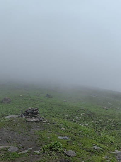 This media file captures a serene and atmospheric scene in a mountainous landscape, likely Dhundi, India, given the context. The primary subjects are a lone hiker walking across a lush, green, grassy slope that disappears into a dense fog. The terrain is uneven, with scattered rocks and patches of mud, indicating a natural, possibly rugged, environment.

In the foreground, a cairn, a man-made pile of stones, stands as a marker or navigational aid. A white arrow is painted on one of the stones, pointing upwards and to the left, suggesting a trail or direction. The presence of this cairn and the arrow hints at a hiking or trekking path.

The weather is overcast and foggy, creating a diffused light and obscuring the distant views. The fog lends an ethereal and somewhat mysterious quality to the image, emphasizing the isolation of the hiker and the vastness of the landscape. The time of day is indeterminate due to the overcast conditions, but the soft light suggests it could be morning or late afternoon.

The overall mood is one of peaceful solitude and exploration. The limited visibility due to the fog could evoke a sense of wonder and anticipation about what lies beyond. There is no discernible text other than the white arrow on the cairn.
