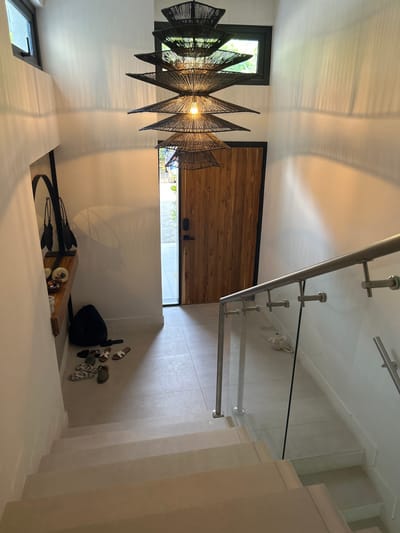 The image shows the interior of a home located at the coordinates 9.658 latitude, -84.673 longitude in the city of Playa Herradura, Costa Rica. A set of stairs leads down to a hallway with a wooden door at the end and a glass railing on the right. The hallway is tiled with light-colored tiles and has a large black, woven lamp hanging from the ceiling. There is a small entryway to the left of the hallway, with a shelf and a backpack. A set of sandals lie on the floor. Light filters in through a window at the top of the hallway. The stair steps appear to be made of a light-colored stone, with a handrail on the left side. The environment is clean and modern, decorated in a minimalist style.