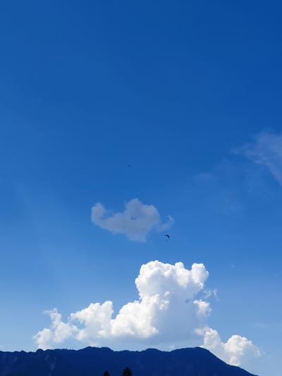 The image depicts a bright, clear blue sky with scattered white clouds. In the distance, a dark mountain range with some sparse vegetation is visible along the horizon. Two paragliders are present in the sky. One paraglider is situated in the upper middle section of the sky, and another is in the lower middle section, closer to the clouds. The lighting suggests it is daytime, with the sun illuminating the scene from an unseen position. No specific buildings or urban features are identifiable, suggesting an outdoor, likely mountainous or hilly, environment. The location is identified as being in or near Levico Terme, Italy.