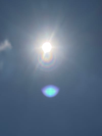 The image shows a bright sun with lens flares against a dark blue sky. There are also some faint, out-of-focus clouds visible in the upper left corner. One lens flare has a rainbow-like effect, and another lower down appears as a bluish-green oval. The image is a photograph taken on a clear day. The location in Tewkesbury, United Kingdom is not discernible from the image.