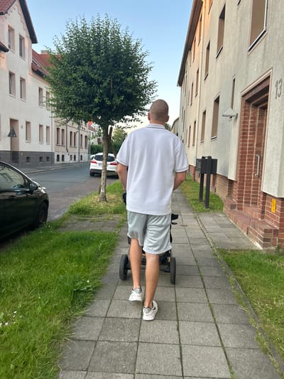 The image shows a person from behind, walking away from the camera on a paved sidewalk. The person is wearing a white polo shirt and light grey athletic shorts, with white athletic shoes. They are pushing a stroller with black wheels. To the left of the sidewalk is a strip of green grass, and beyond that, a dark-colored car is parked on the side of a street. Across the street, there are residential buildings with light-colored facades and red-tiled roofs. A slender tree with green foliage stands between the sidewalk and the street. To the right of the sidewalk, there is another building with a light-colored facade, a brick entrance, and a mailbox. The scene is illuminated by the soft light of dusk. The location is identified as Schöningen, Germany.