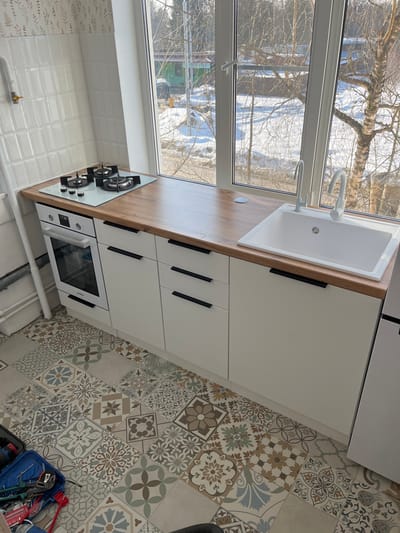 A well-lit kitchen in the city of Нарынка, Russia, features a modern interior design. The focal point is a light-toned wooden countertop extending along a large white-framed window. Integrated into the countertop are a white three-burner gas cooktop on the left and a white rectangular sink with a white main faucet and a smaller secondary faucet on the right. Below the cooktop is a built-in white oven. The base cabinetry consists of white, handleless flat-panel units with long black horizontal bar handles. The wall behind the cooktop is tiled with white rectangular subway tiles, above which is a subtly patterned wallpaper featuring leafy motifs. A white vertical pipe runs along the left wall. The floor is covered in a mosaic of patterned ceramic tiles in various muted colors including brown, grey, blue, and green. In the lower-left foreground, a blue toolbox filled with various hand tools, including pliers and wrenches, is visible on the floor.
Through the multi-pane window, an exterior winter scene is visible. The ground is covered in snow, and numerous deciduous trees, including a prominent birch tree on the right, are bare. Distant evergreen trees are also present. Pathways or roads are discernible through the snow. In the mid-ground, a green building and a smaller yellow structure can be seen. The scene is illuminated by bright daylight, casting no discernible shadows. No individuals or specific actions are present in either the interior or exterior view.