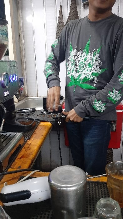The scene depicts an individual preparing coffee within a commercial establishment in RawangItiek, Indonesia. A person, partially visible from the chest down, wears a grey long-sleeve shirt adorned with large green and white abstract leaf designs on the chest and sleeves, paired with blue jeans. The individual is actively engaged in the coffee preparation process, using their right hand to tamp ground coffee within a portafilter, which is held steady by their left hand. This action takes place on a dark tamping mat positioned on a light wooden countertop. To the left, a silver espresso machine with blue digital displays indicating "00" is visible, featuring a clear hopper containing coffee beans. In the foreground, on the dark counter mat, an upside-down silver metallic cup, a white electrical appliance with a black handle, a clear glass, and an amber-colored glass are present. The background features a white paneled wall, a visible sink, and a red cooler or storage container to the right of the person. A woven brown decorative item hangs on the background wall.