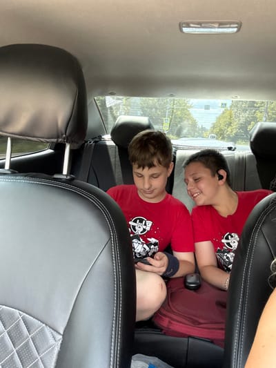 In the image, two young boys are seated in the back of a car. Both boys are wearing red t-shirts with a graphic print. The boy in the foreground, who is partially obscured by the seat in front, appears to be resting with his eyes closed. The boy behind him is looking towards the camera with a smile and has earbuds in. The car interior features black leather seats with white stitching. Through the windows, trees and a road with traffic signs are visible, suggesting a daytime setting on a clear day. One of the signs appears to be a yield sign, indicating this is likely a road in Russia, specifically Goryachy Klyuch, given the context. There is no visible text within the image itself.