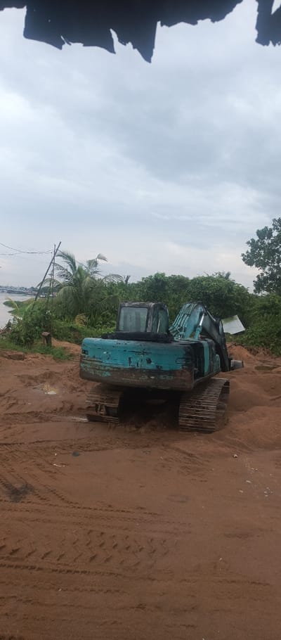 The image displays an outdoor scene along the Kapuas River in Pontianak, Indonesia, under an overcast sky. In the midground, a stationary, heavily weathered teal-colored tracked excavator is positioned on an elevated mound of reddish-brown sand and dirt. The excavator's cab and retracted boom are visible; no operator is present. The foreground consists of reddish-brown sand and mud, marked by distinct excavator track impressions and tire tracks, with several small water puddles present. The background to the left reveals a section of the Kapuas River with lush green tropical vegetation, including palm trees, lining its banks. A portion of the Kapuas I Bridge, a prominent steel truss bridge, is visible spanning the river in the distance. Utility poles and overhead wires are present on the left. The background to the right features dense green foliage and a small, partially visible light-colored structure. The top edge of the image is obscured by an irregular dark shape. No individuals are visible in the scene.