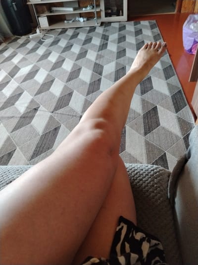 A person's legs are crossed, with the right leg on top of the left. The right foot is visible, with painted toenails. The person is sitting on a couch with a textured gray fabric. Behind the couch is a geometric patterned rug in shades of gray and white. In the background, there is a shelf unit with various items on it, and a black suitcase. The lighting suggests it is daytime. The scene appears to be indoors, in a living room or similar space.