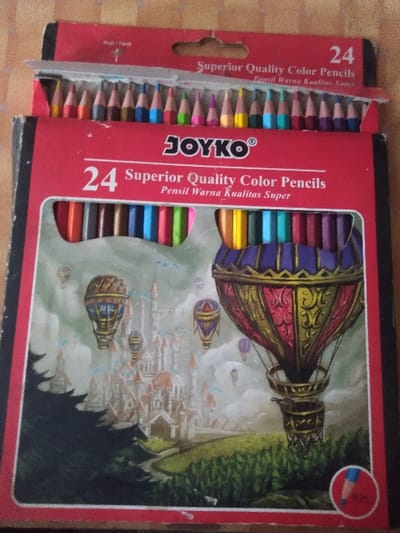 The image displays an open red cardboard box of "JOYKO" brand "24 Superior Quality Color Pencils" (also labeled "Pencil Warna Kualitas Super" in Indonesian). The top flap is pulled open, revealing a row of 24 sharpened colored pencils with visible tips, arranged horizontally. The box's main front panel features an illustration of a fantasy landscape with multiple hot air balloons of various sizes and colors floating over a large, multi-spired castle nestled among green hills and dark trees, beneath a cloudy sky. The lower right corner of the illustration includes a small circular icon depicting a blue colored pencil. The box rests on a light brown, textured surface, likely wood, visible in the background and foreground. No people, actions, or interactions are depicted. There is no discernible information within the image to indicate its specific location in Жарсуат, Kazakhstan, or any other geographical area.