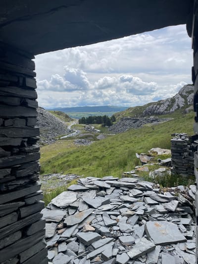 The image captures a view through an opening framed by rough-hewn stone, likely slate, into a landscape characterized by rolling green hills, rocky outcrops, and scattered trees. In the foreground, a pile of fragmented grey slate pieces fills the bottom of the frame, suggesting a former quarry or construction site. A path winds through the green landscape, with several figures walking along it in the mid-ground. Beyond the path, the terrain becomes more rugged with steep, rocky slopes and further piles of discarded stone. The sky above is filled with a mix of white and grey clouds, indicating a potentially overcast day. The scene appears to be an outdoor industrial or post-industrial landscape, possibly within or near Tanygrisiau, United Kingdom, given the characteristic slate formations and hilly terrain.