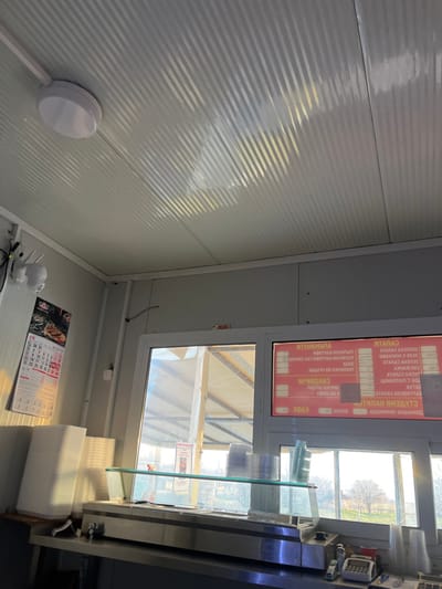 The image depicts the interior of a commercial establishment in Petkovo, Bulgaria, characterized by a utilitarian design. The ceiling is composed of white, ribbed paneling, featuring a single circular white LED light fixture positioned in the upper left quadrant. Sunlight reflects off the ceiling, creating a pattern of diagonal light and shadow. The walls are constructed from light gray panels.
On the left wall, a white monthly calendar for "ЮЛИ" (July) hangs, displaying dates and possibly an image at the top. Below the calendar, on a stainless steel counter, a tall stack of white rectangular disposable food containers is present. Above the calendar, a white security camera is mounted on the wall, with associated wiring visible.
A prominent window unit dominates the center and right side of the image, offering a view to the exterior. The upper right pane of this window displays a digital menu board with a red background and white Cyrillic text, listing various food and beverage items such as "САЛАТА" (salad) and "КАФЕ" (coffee). Directly in front of the window, a clear glass display shelf rests on the stainless steel counter, holding stacks of clear plastic cups or containers. To the far right on the counter, several smaller items, including a white card reader and an electronic scale, are discernible.
Through the window, an outdoor structure with a light-colored, possibly fabric or tarp, roof supported by metal beams is visible immediately outside. Beyond this structure, a distant treeline of bare trees suggests an open, possibly field-like, landscape under bright, direct sunlight. No individuals are present within the frame.