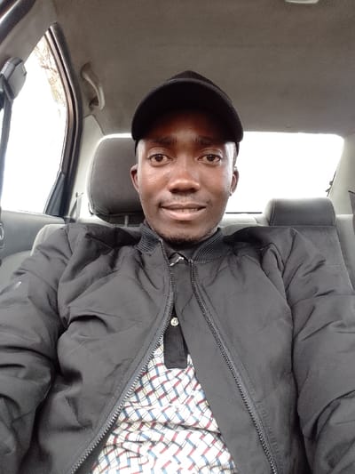A single adult male is seated within the interior of a vehicle, located in Matapato North ward, Kenya. The individual, with a dark complexion, wears a black baseball cap, a black quilted or puffer jacket, and a patterned polo shirt featuring red, blue, and white plaid or geometric designs. He is positioned facing the camera, exhibiting a slight smile. The vehicle's interior includes grey fabric headrests and seating surfaces. Bright, undifferentiated light is visible through the vehicle's windows, suggesting an outdoor environment with overcast conditions or significant sky brightness. No other persons or significant objects are visible within the frame beyond the individual and vehicle interior elements.
