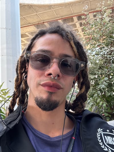 A medium shot depicts a male with dreadlocks and sunglasses. He has a nose ring and a short beard. He is wearing a navy blue t-shirt under a black zip-up jacket. Headphones are visible. Behind him are plants and what appears to be a beige architectural structure. While the location is specified as Montaña Roja, Spain, this cannot be confirmed from the visual information alone.