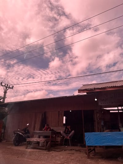 The image depicts an outdoor scene in Buengbakjok, Indonesia, under a pink-tinged sky filled with clouds. In the foreground, a wooden building with a corrugated metal roof stands. To the left of the building, a motorcycle is parked. In front of the building, two men are seated. One man, wearing a red shirt, sits at a wooden table, leaning on it with his arm. Another man, wearing a dark shirt and grey pants, sits on a chair to the right, looking at his phone. A blue tarp covers a table or structure to the far right. Power lines stretch across the sky, supported by a utility pole visible on the left. The ground appears to be a dirt and gravel surface.