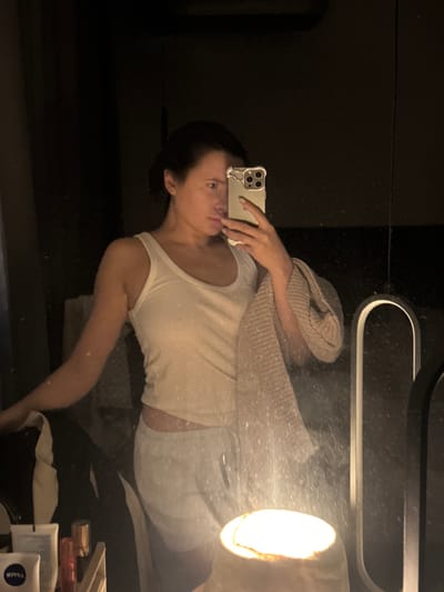 An adult female is depicted in a mirror selfie within a dimly lit indoor environment, identified as the city of Селекционер-2 Снт, Russia. She wears a white tank top and light-colored sweatpants. Her dark hair is pulled back. She holds a smartphone with a light-colored case featuring ear-like protrusions in her right hand, positioned to capture her reflection. A light-colored, ribbed textile, possibly a towel or scarf, hangs over her left forearm. The mirror surface exhibits visible particles and smudges. In the lower central foreground, a bright, warm-toned cylindrical light source illuminates the scene. A partial reflection of an arched light fixture is visible on the right. To the lower left, on a reflective surface, several personal care products are arranged, including a white tube labeled 'NIVEA'. The background beyond the reflection is largely dark.