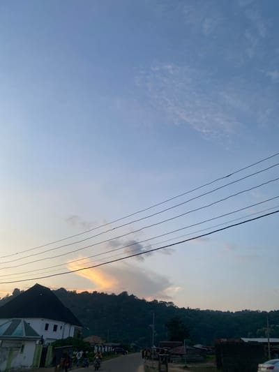 This image, captured in Enwan, Nigeria, depicts a late afternoon or early evening scene. The upper two-thirds of the frame are dominated by a pale blue sky, transitioning to a lighter, warmer hue near the horizon. Sparse, wispy white clouds are scattered across the sky, with a notable, elongated cloud formation above the left portion of the treeline exhibiting a distinct orange-yellow glow on its underside, indicative of a setting sun. Five dark, parallel power lines stretch horizontally across the middle of the sky.
The lower third of the image features a landscape comprising buildings, a road, and densely forested hills. In the lower-left foreground stands a prominent multi-story building with white walls and a dark, possibly black or dark grey, hip roof. To its immediate right, a smaller building with a light green-tiled roof and white walls is visible. Behind these structures, a range of dark green, tree-covered hills or mountains forms the backdrop, rising from the mid-ground.
A road traverses the lower section of the image, where several individuals and vehicles are present. At least two motorcycles, each with a rider, are visible on the left side of the road, moving towards the right. Other people are standing or walking along the roadside. A dark-colored SUV is parked on the right side of the road, and the front portion of a white car is partially visible at the extreme right edge of the frame. The overall lighting is soft and warm, characteristic of dusk.