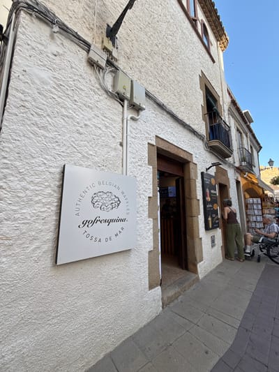This is a low-angle, wide-shot photograph of a textured, off-white building facade with a clear blue sky above. The building appears to be an older establishment, possibly in a European coastal town, given the architectural style and the visible sign.
In the foreground, a white sign with black text and an illustration of a waffle is mounted on the wall. The text on the sign reads, "AUTHENTIC BELGIAN WAFFLES" in an arc around a stylized waffle graphic, and "gofresquina TOSSA DE MAR" in a curved line below.
Above and to the right of the sign, a section of the building reveals a recessed window or doorway leading into a shop. Inside, glimpses of shelves and potentially food items are visible. Further up the facade, a person is lying down on the exterior of the building, seemingly resting or sunbathing. Their legs are extended outward, wearing olive green pants, and they are lying on their stomach, facing downwards. Above them, another person, wearing a striped shirt and shorts, stands near a bicycle with a basket, suggesting a casual, daytime setting.
The scene suggests a warm, sunny day. The presence of the waffle shop sign and the relaxed posture of the people indicate a leisurely atmosphere, likely in a tourist-oriented area. Electrical conduits and junction boxes are also visible on the wall, suggesting modern utilities integrated into the older structure. The overall impression is one of a relaxed, sun-drenched day in a charming, perhaps historic, urban environment.