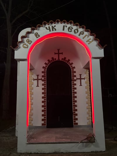 The image shows a small, white, rectangular structure with a rounded arch at the top, likely a roadside shrine or a small religious building. The structure is adorned with a red neon light that traces the archway and the top horizontal line. The inscription "СВ. ГЕОРГИ" is visible on the left side of the arch, suggesting it is dedicated to Saint George. The interior of the structure is dark, with crosses painted on the back wall. The front of the structure has an open doorway. The surrounding environment is dark, suggesting it is nighttime. The location is Sungurlare, Bulgaria.