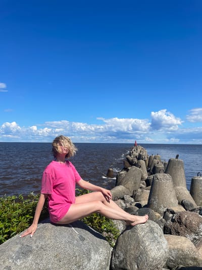A young woman with blonde hair, wearing a bright pink t-shirt, sits on a large grey rock on the shore of the Beloye Lake in Belozersk, Russia. Her legs are crossed, and she is looking towards the water, with her hair partially obscuring her face. In the background, a long breakwater made of concrete tetrapods extends into the dark blue water under a bright blue sky with fluffy white clouds. The scene is set during the daytime, indicated by the strong sunlight and clear weather.