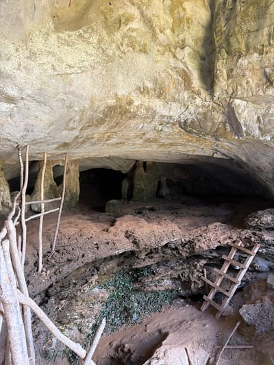 The image shows the entrance to a dark cave or tunnel carved into a rocky hillside in Ban Phon Sai, Laos. The cave opening is roughly oval and appears to lead into a deep, dark interior. The rock face is textured and appears weathered, with some lighter, smoother sections and rougher, darker areas.

A makeshift wooden ladder is propped against the rock face on the right side of the cave entrance, suggesting this is a route for exploration or access. Another, more rudimentary ladder constructed from branches and logs spans across the opening, likely to provide a pathway into the cave. These structures are rough and appear to be constructed from natural materials found in the environment.

The ground leading up to the cave entrance is composed of dirt and loose rocks, with some sparse green vegetation growing on the hillside to the right of the entrance. The overall impression is one of an ancient or natural site that has been made accessible through rudimentary means. The lighting suggests it is daytime, with shadows indicating the sun is present but perhaps not directly overhead. There is no visible text in the image.
