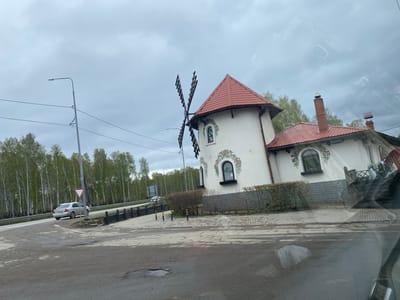 The image shows a small windmill-shaped building with white walls and a red tiled roof, located at a roadside intersection. The windmill has four black sails and decorative flower paintings around the arched windows. A single-story extension adjoins the windmill, also with a red tiled roof and matching floral decor. A brick chimney rises from this section. A low brick wall and a trimmed hedge surround the building.
A road runs alongside the building, with a silver car making a turn at the intersection. A streetlight stands at the corner. In the background, a line of trees is visible. The sky is overcast. The image appears to be taken from inside a vehicle, as part of the dashboard is visible in the foreground.
The location is Молодёжный, Russia.