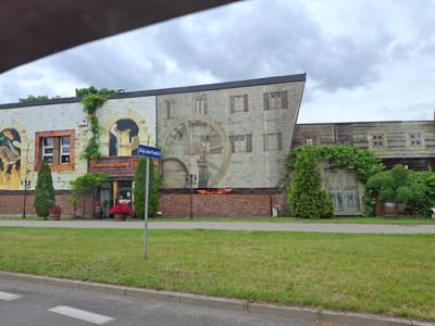 A building with a mural depicting a historical scene is the main subject. The mural features a large water wheel, windows with shutters, and a rustic brick facade. In front of the building, there is a grassy area with a sidewalk and a street. Several evergreen trees and potted plants with red flowers add greenery and color to the scene. A street sign reading "Aleja Jana Pawła II" is visible on the left side of the building. A sign above the entrance reads "Pomarańczowy Folwark RESTAURACJA WIK." Another sign on the brick wall appears to say "MOLINO". The sky is overcast with clouds, suggesting it might be a cloudy day. The scene appears to be a restaurant or commercial establishment in Pila, Poland. No people are visible in the image.