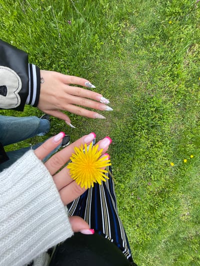 The image depicts a first-person view from above, looking down at two hands and a grassy field. One hand holds a yellow dandelion. The person is sitting down, wearing blue jeans, a white sweater and a black varsity style jacket. The grassy field contains some yellow dandelions. The image does not provide information about the city of Barkach, Bulgaria.