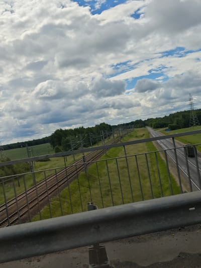 The image shows a view from a bridge overlooking a railway and a highway. The sky is filled with dramatic clouds. On the left side of the frame, a set of train tracks runs parallel to the highway. On the right, the highway curves into the distance, with a single truck visible. A dense line of trees borders the landscape beyond the tracks and highway. Overhead power lines are supported by tall metal towers, one of which is prominent on the right side of the image. The foreground of the image is dominated by a metal railing and the surface of the bridge. The scene appears to be in a rural or semi-rural area, possibly on the outskirts of a town or village. The location is described as Malewiccki Selski Sovet, Belarus.
