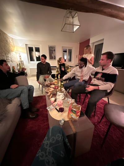 A gathering of six people is centered around a low coffee table laden with food and beverages. Individuals are seated on chairs, a sofa, and even perched on the edge of the table itself. One person is pouring a drink. The setting appears to be a cozy living room, illuminated by a warm lamp and natural light filtering through windows. A decorative ceiling fixture and artwork on the walls add character to the space.
