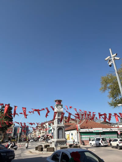 This image shows the city center of Iznik, Turkey, on a sunny day. The focus is a white stone clock tower, decorated with Turkish flags and illuminated against a bright blue sky. Overhead are suspended strings of additional flags that span the main street, creating a festive atmosphere. Vehicles are parked on the side of the road, and a street light with multiple security cameras is visible in the upper left corner. Trees frame the scene at the top, while storefronts and commercial buildings line the street.
